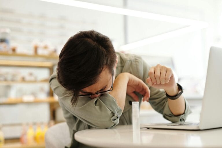 - Labasa An adult man sneezes into his arm near a laptop in a cafe, illustrating illness prevention.