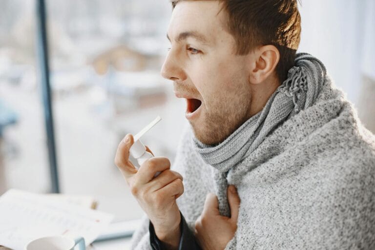 - Labasa An adult man uses a throat spray, wrapped in a blanket, appearing unwell indoors.